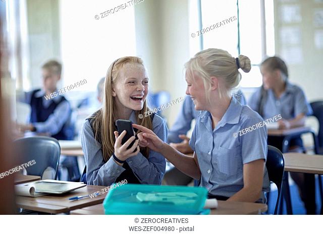 Two happy schoolgirls in classroom with cell phone