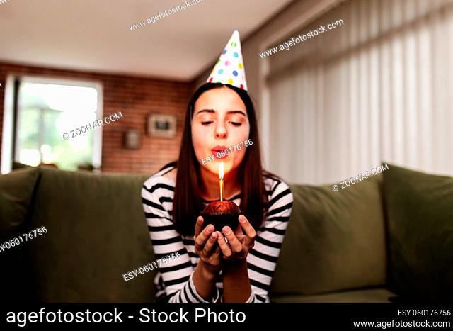 Birthday girl brunette in cardboard hat sits on the couch or sofa and blows out burning candle on cupcake. Woman holding birthday cake in her hands