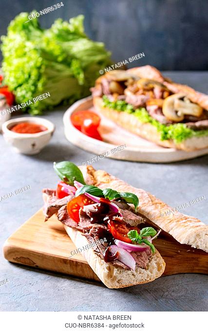Beef baguette sandwich with tomatoes, basil, red onion served on ceramic plate with ingredients above over grey blue table