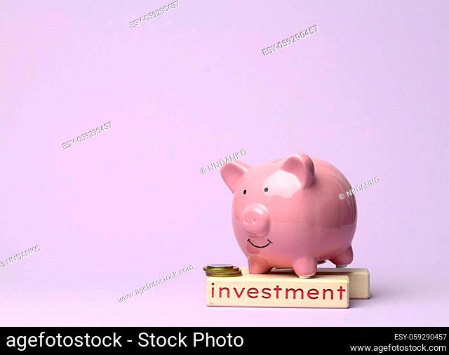 ceramic pink piggy bank and wooden cube with the inscription investment on a lilac background, concept of savings and long-term financial planning