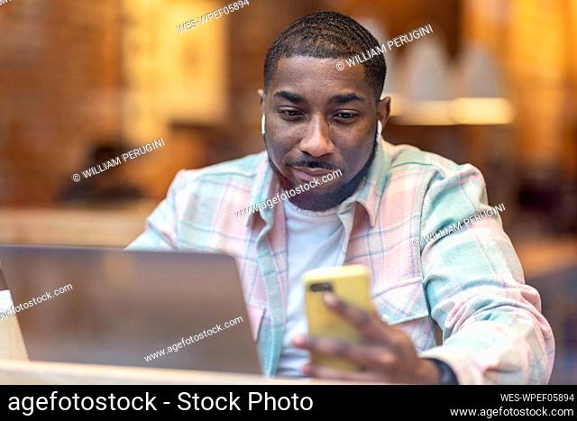 Man with laptop listening music through wireless in-ear headphones using mobile phone in cafe