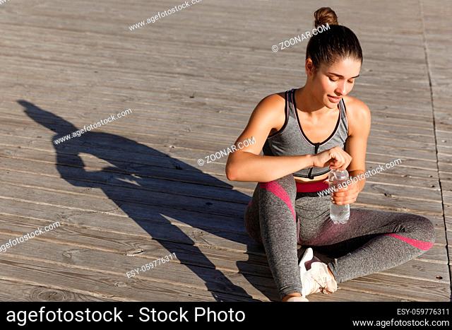 Outdoor shot of attractive fitness woman having a break after jogging, sitting on floor and drinking water, working out near the sea