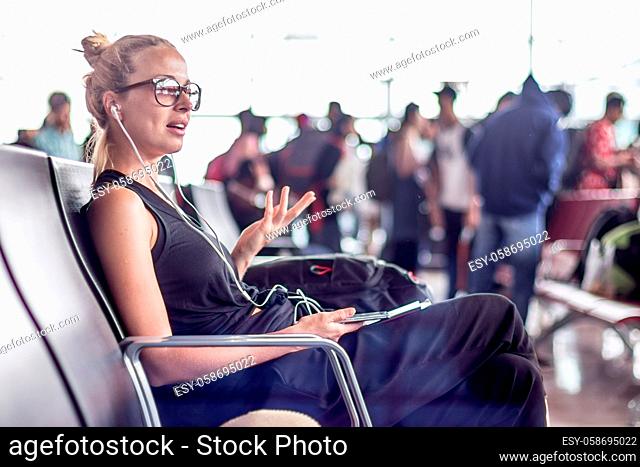Casual sporty young blond female traveler talking on her cell phone while waiting to board a plane at the departure gates at the asian airport terminal