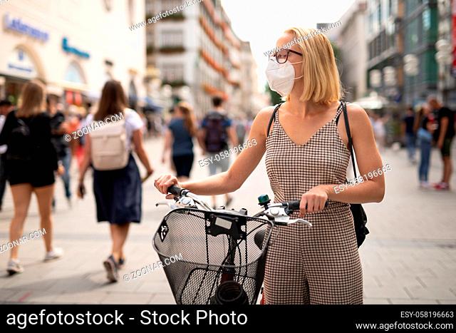 Woman walking by her bicycle on pedestrian city street wearing medical face mask in public to prevent spreading of corona virus