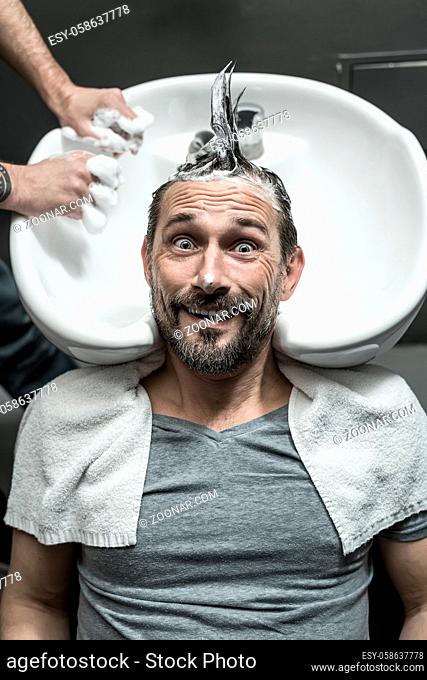 Grimacing man with a beard lies on the white sink in the barbershop. He has the lathered head with the punk hairstyle. Guy wears a gray T-shirt with a white...