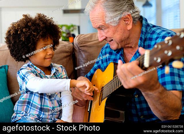 Multiracial grandfather holding grandson's hand and teaching him to play guitar on sofa at home