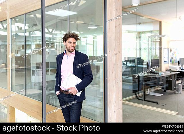 Male entrepreneur holding document while leaning on glass wall in factory