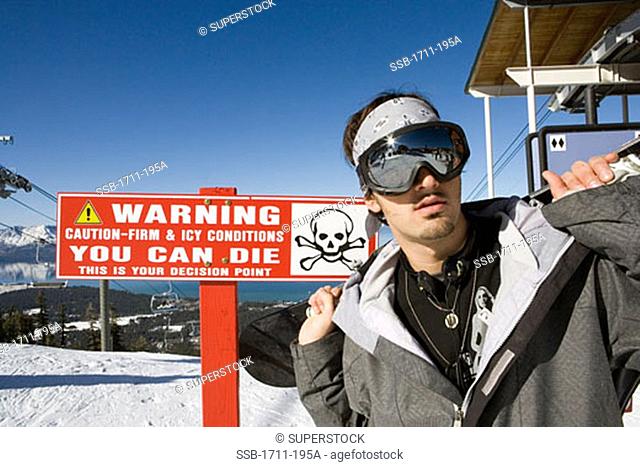 Young man carrying a snowboard in front of a warning sign