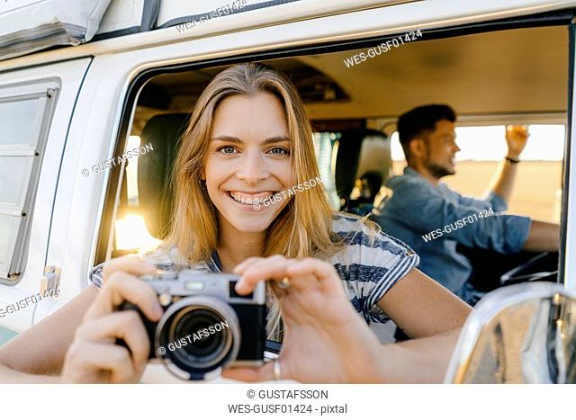 Portrait of happy woman with camera leaning out of window of a camper van with man driving