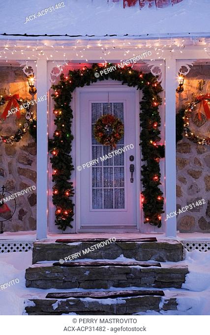 Illuminated main entrance door of a two-storied Canadiana style residential home decorated with a Christmas wreath and lights at dusk, Laval, Quebec, Canada