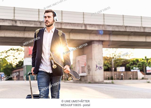 Young man on the move with skateboard, rolling suitcase and headphones