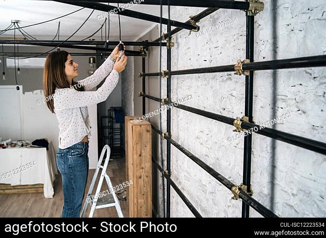 Mid adult woman on step ladder hanging lightbulb in her new shop