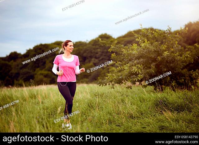 Fit middle-aged woman walking through a rural field listening to music on her mobile phone using earbuds looking to the side with a happy smile in a healthy...