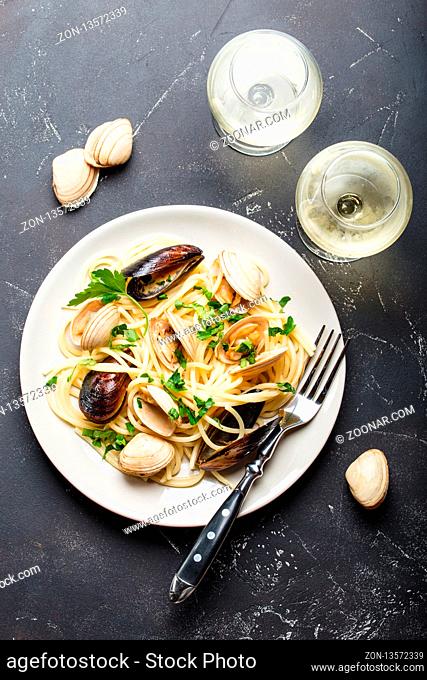 Spaghetti vongole, Italian seafood pasta with clams and mussels, in plate with herbs and two glasses of white wine on rustic stone background