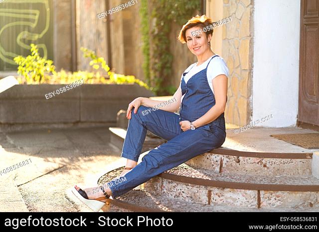 Adult Pregnant Caucasian Woman In A Blue Jumpsuit Rests Sitting On The Porch Of The House Outside On A Summer Day In Modern European City Anticipation Concept