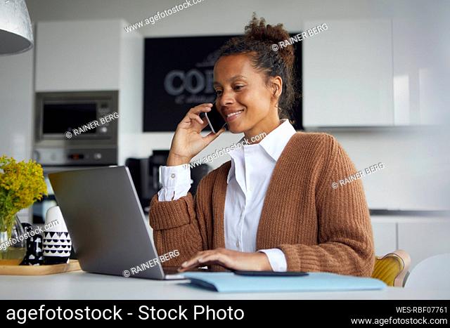 Smiling businesswoman on the phone sitting in kitchen working on laptop