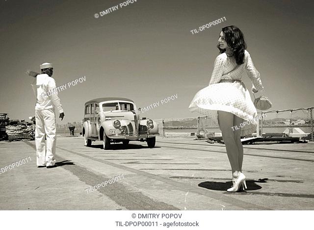 A model wearing a short white dress near a sailor and vintage car