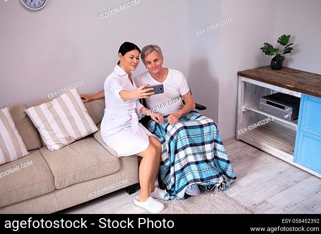 Young nurse taking selfie with patient. Sweet asian nurse in white clothes taking pictures with aged disabled man in wheelchair