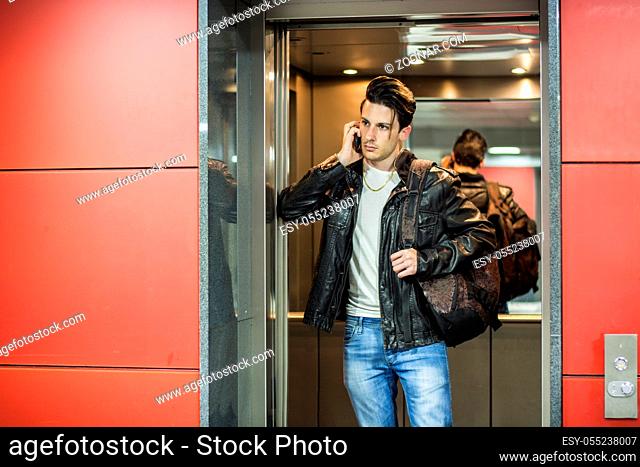 Handsome young man leaning against mirror inside an elevator or lift, calling someone on cell phone