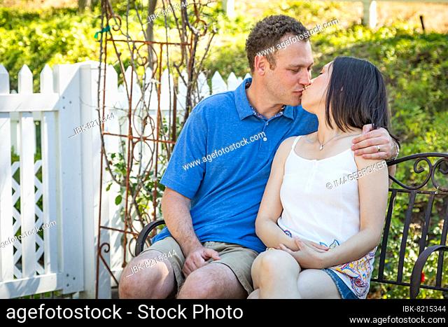 Young caucasian man and chinese woman sitting on bench kissing