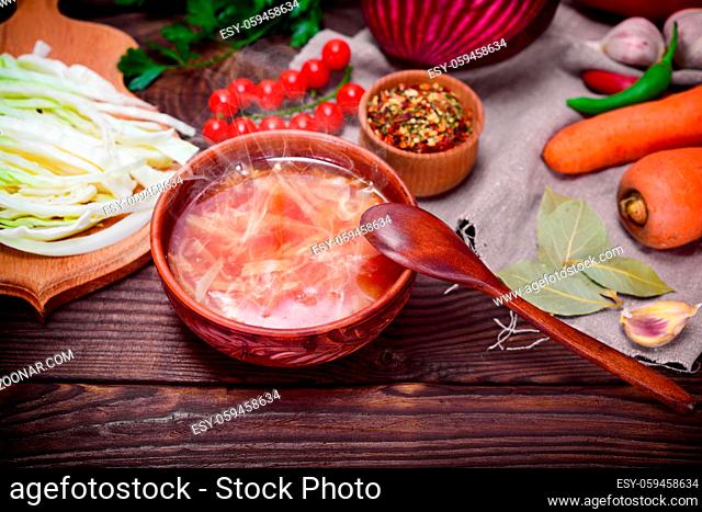 Traditional Ukrainian borsch in a round brown plate in the middle of vegetables and ingredients on a wooden table