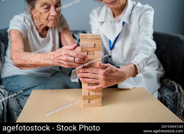 Dementia therapy in playful way, training fingers and fine motor skills, build wooden blocks into tower, playing Jenga. Senior woman 90 years old and doctor...