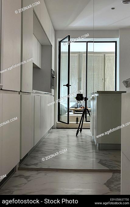 Reflection in the mirror of the modern kitchen zone and a glass door entrance to the glowing room with tall windows with curtains
