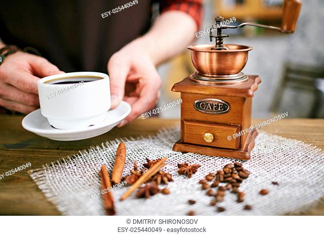 Wooden coffee grinder, coffee beans, star anise, cinnamon sticks on napkin and cup of black coffee in barista hands