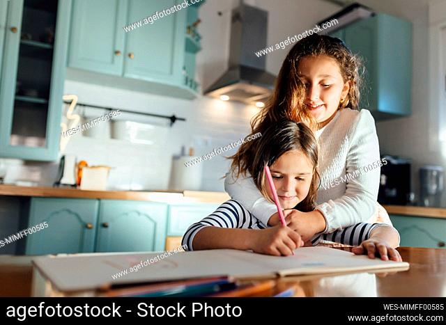 Girl coloring on paper while sister hugging from behind in kitchen