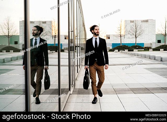 Young businessman with briefcase looking away while walking by glass wall