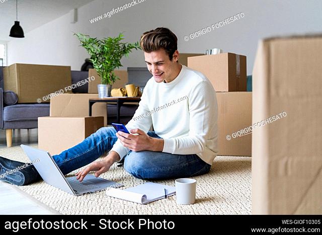 Smiling man with laptop using smart phone while sitting in living room of new home