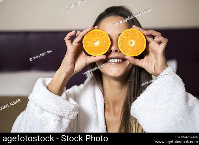 Pretty young brunette woman in white morning gown sitting in hotel room and holding slices of orange near eyes