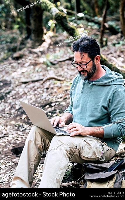 adult caucasian man sitting on the ground in the forest do a online work connection with laptop computer - internet wireless smart working job digital nomad...