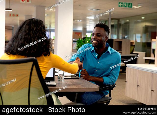 Smiling african american businessman shaking hands with biracial businesswoman sitting at desk