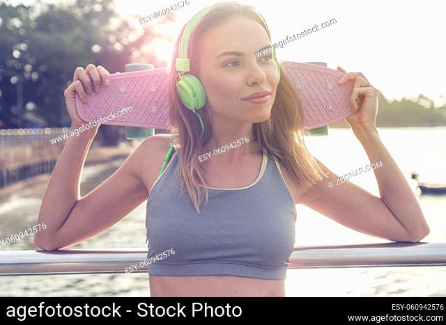 Young pretty blonde fitness female wearing bright green headphones listening music and holding pink skateboard while standing on the pier near the sea