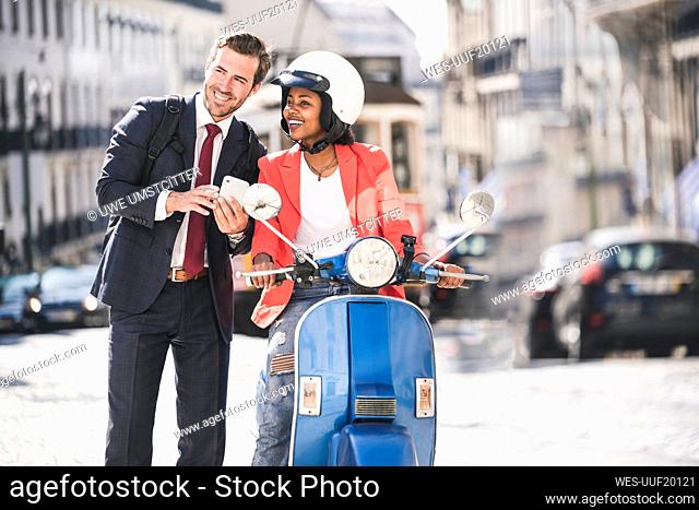 Happy young business couple with cell phone and motor scooter in the city, Lisbon, Portugal