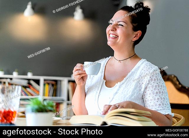 Cheerful young woman looking away while sitting with coffee and book in cafe