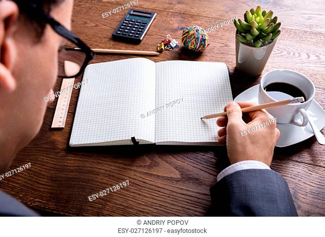 High Angle View Of Businessman Hand With Pencil On Notebook At Wooden Desk In Office