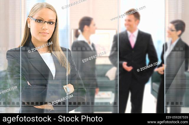 Confident young businesswoman standing in office behind glass wall, businesspeople talking in background