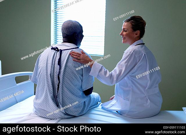 Diverse female doctor and male patient sitting on hospital bed smiling to each other