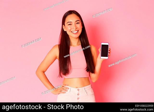 Technology, communication and online lifestyle concept. Gorgeous trendy asian girl standing over pink background with mobile phone