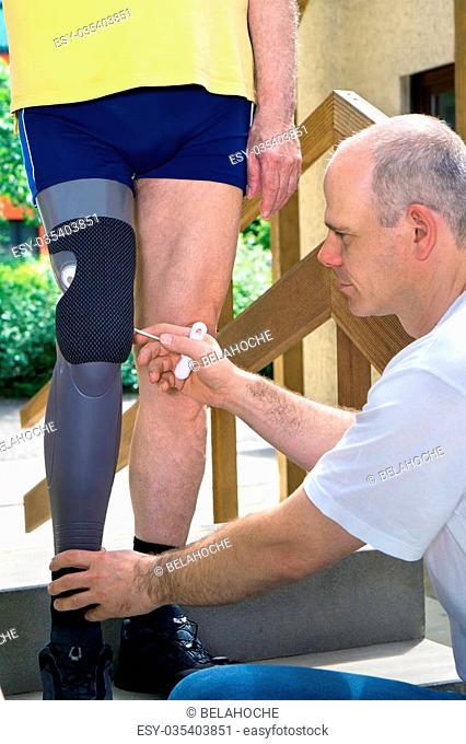 Physiotherapist adjusting gray and black prosthetic leg for unidentifiable male patient in blue shorts and yellow shirt