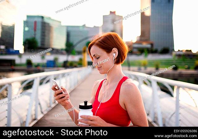 A young woman resting after doing exercise on bridge outdoors in city, using smartphone