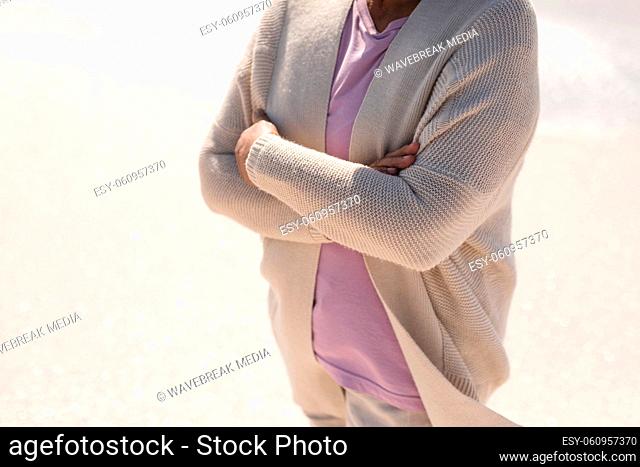 Midsection of senior biracial woman with arms crossed standing at beach on sunny day