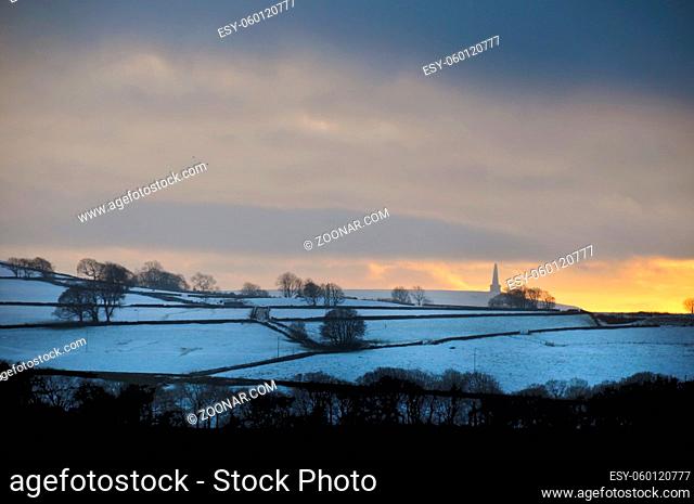 glowing orange sunset in a cloudy twilight winter sky with snow covered fields with stoodley pike monument in the distance in calderdale west yorkshire