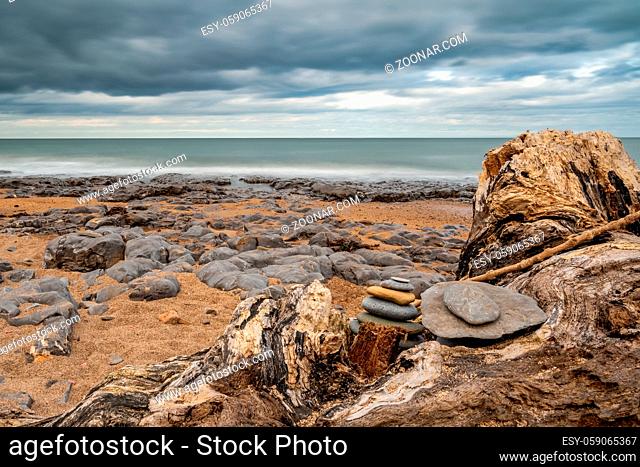 A stone pile on a tree trunk, under a dramatic sky on a stony beach, seen at Cocklawburn Beach near Berwick-upon-Tweed in Northumberland, England, UK