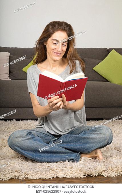 smiling happy brunette woman with grey shirt and blue jeans trousers, reading red book sitting on carpet indoor home