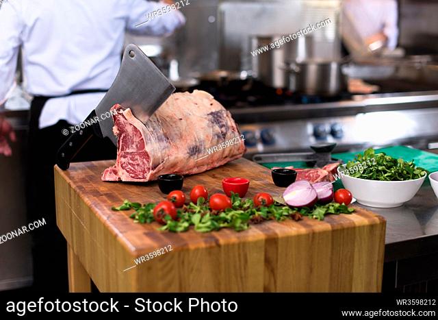 chef using ax while cutting big piece of beef on wooden board in restaurant kitchen