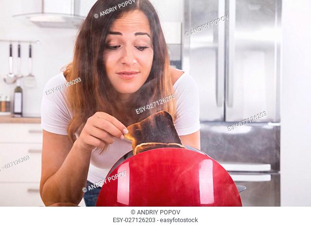 Young Woman Taking Slice Of Burnt Toast Out Of The Toaster In Kitchen