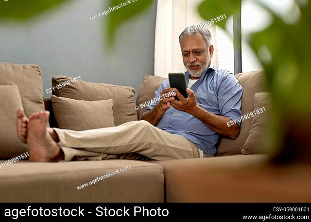 A SENIOR ADULT MAN SITTING COMFORTABLY ON SOFA AND USING MOBILE PHONE